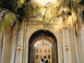 Bride and groom embrace on church steps under palm trees. Wooden doors in the background, warm sunlight, romantic and elegant setting.