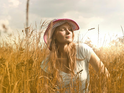 Woman in white shirt and striped hat sits in a sunny wheat field, eyes closed, peaceful expression, golden grass and cloudy sky.