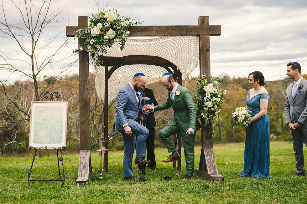 Gay Jewish wedding photo of two grooms smashing glass underfoot as the ceremony concludes, under a chuppah and next to their ketubah.