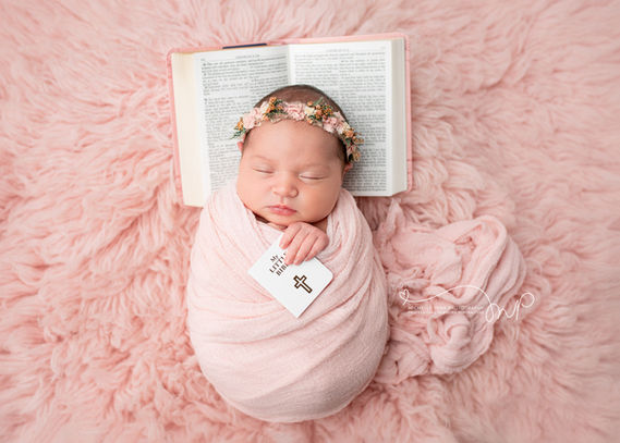 Baby photography using a pink rug and pink wrap. She is posed on a bible and is also holding a smaller bible.