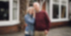 An elderly couple smiles outside a red brick house with large windows. The woman wears a blue sweater and scarf, the man a red pullover.