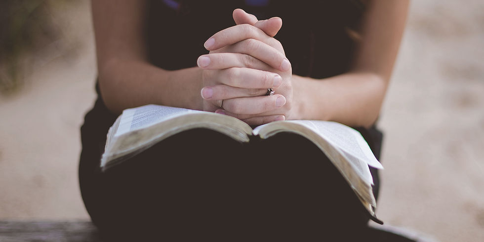 Hands clasped in prayer over an open book on a wooden bench. The background is blurred, creating a serene, contemplative mood.