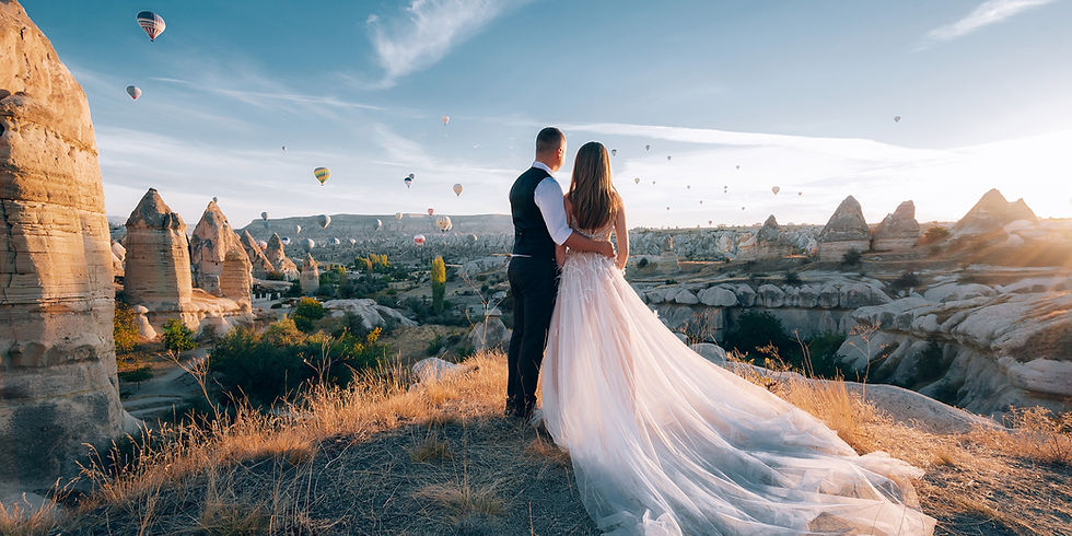 A couple embraces, gazing at a landscape with hot air balloons over rocky formations at sunset. Bride in a flowing white dress; romantic mood.