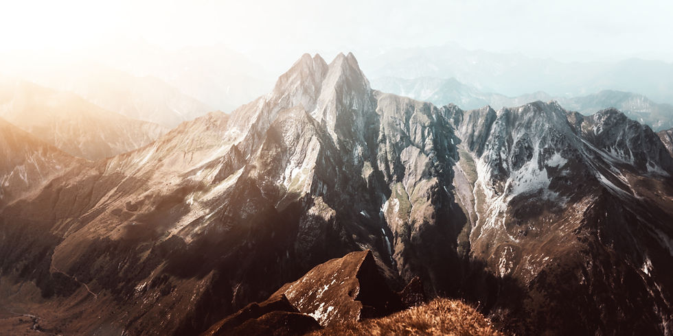 Mountain range with sharp peaks under a bright sky. Warm sunlight illuminates rocky terrain with patches of snow, creating a serene mood.