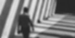Silhouetted person walking through shadowed, striped corridor; light patterns create a dramatic, moody atmosphere. Black and white image.