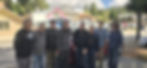 Eight men standing shoulder to shoulder in front of a parked car in a small mountain town on arrival day.