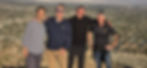 Four men standing arm in arm on a rocky ridge, with a desert valley of houses spread out far below them.