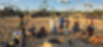 About a dozen men in a circle of camp chairs around an unlit fire ring on the beach, with palm trees and hillside houses behind them.