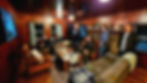 Eight men gathered in a warmly lit wood-panelled cabin living room in the evening, some seated and some standing.
