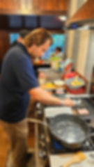A man cooking bacon and eggs at a cabin stove while another man works in the kitchen behind him.