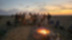 A large group of men standing and seated behind a lit beach fire ring at dusk, with an orange sunset over the water behind them.