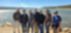 Seven men standing together on a lakeshore, with forested hills and open water behind them.
