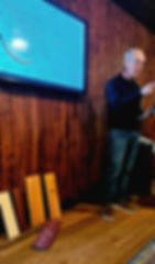 A man speaking beside a screen reading From Rough Lumber to Refined Purpose, with finished wooden boards leaning against the wall below it.