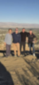 Four men standing together at a desert overlook, with a wide valley and a mountain range behind them.