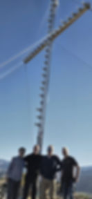 Four men standing at the base of a tall guyed antenna tower at the summit of a hike, under a clear blue sky.
