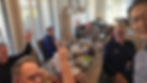 Six men around a dining table waving at the camera during a group selfie, with plates of food in front of them.