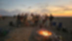 A large group of men gathered behind a burning beach fire ring at sunset, one man with his arm raised.