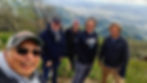 Five men smiling in a group selfie partway up a green mountain trail, with a valley visible far below.
