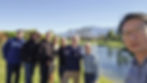 Eight men standing together on grass beside a golf course pond, with a mountain in the distance.