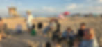 Men seated in camp chairs on a beach listening to a man in a straw hat speaking, with a lifeguard tower behind them.