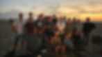 More than twenty men standing close together on the beach at dusk for a group photograph, with the ocean and a fading sunset behind them.