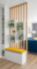 Modern living room divider with white shelves, potted plants, and a yellow bench, beside a blue sofa and floor lamp.