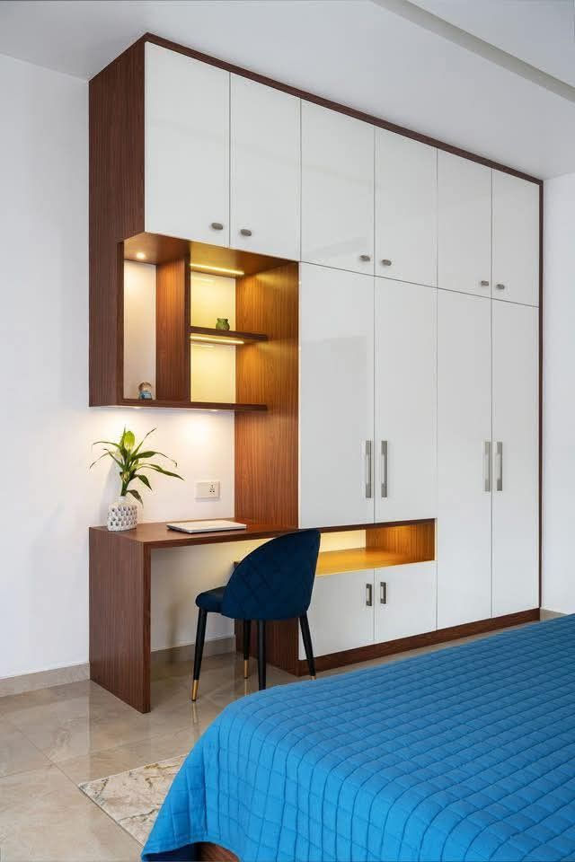 Modern bedroom with white and wood built-ins, a small desk and blue chair, warm shelf lighting, and a blue quilted bedspread.
