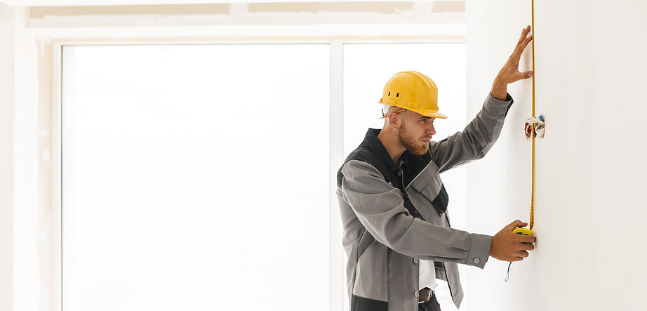 young-builder-work-clothes-yellow-hardhat-using-measuring-tape-repairing-flat-with-big-win