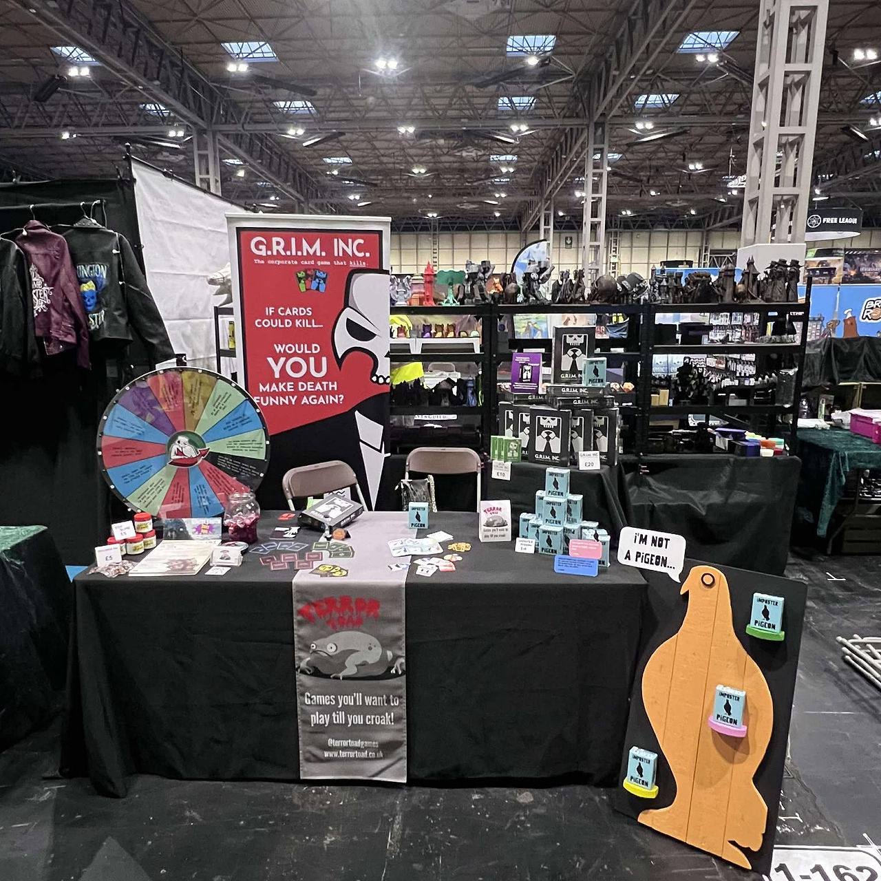 Our stall from UKGE 2024. Copies of Imposter Pigeon, GRIM INC, our spinning wheel, banner, and Toad table runner all on show