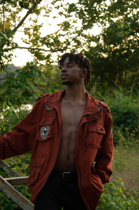 Young man in red jacket standing outside amidst lush green foliage.