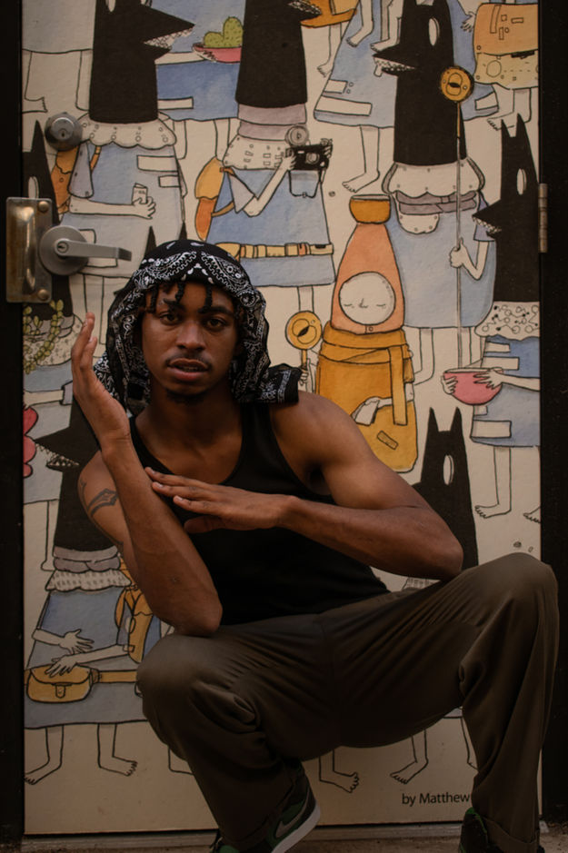 Young man poses with hand gestures, wearing bandana, dark tank top, mural background