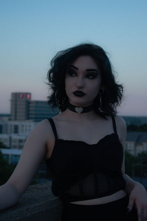 Woman in black outfit, choker, dark makeup looks out against city background.