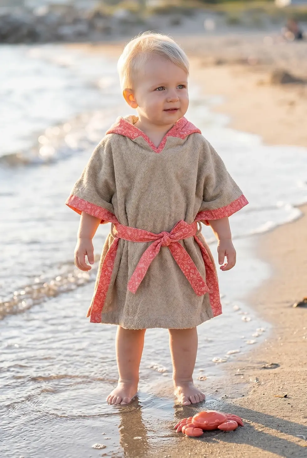 Miniaturbild: Blondes Kleinkind steht barfuß am Strand im personalisierten Badeponcho „Korallenbucht“ von Küstenfieber®.