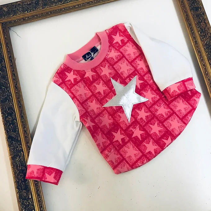 Miniaturbild: Detailaufnahme eines handgemachten Mädchenpullovers mit pinkem Sternenmuster, weißen Ärmeln und appliziertem Silberstern.