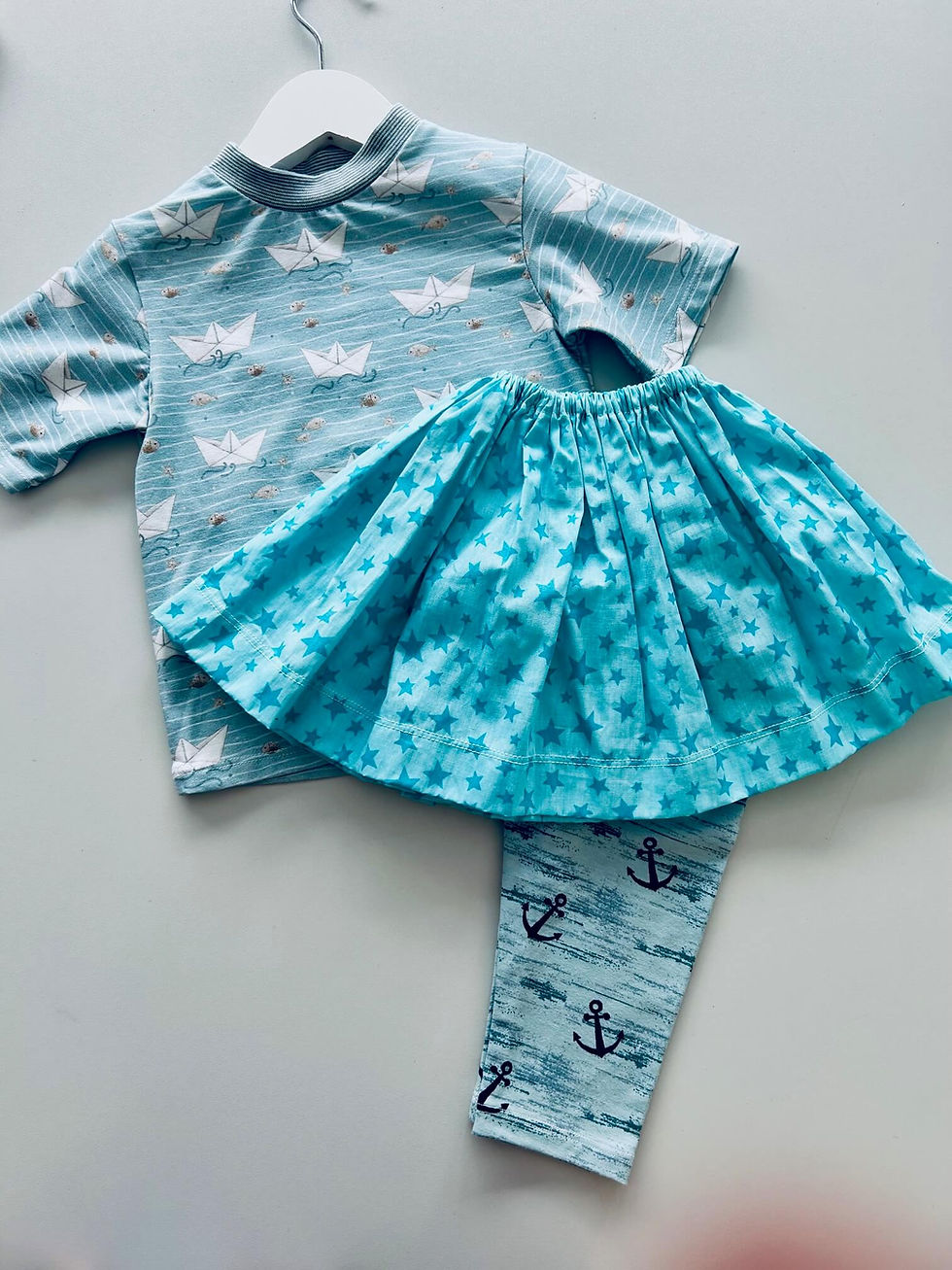 Niedliches Mädchen-Outfit im maritimen Stil mit Papierschiffchen-Shirt, türkisfarbenem Sternenrock und Anker-Leggings.