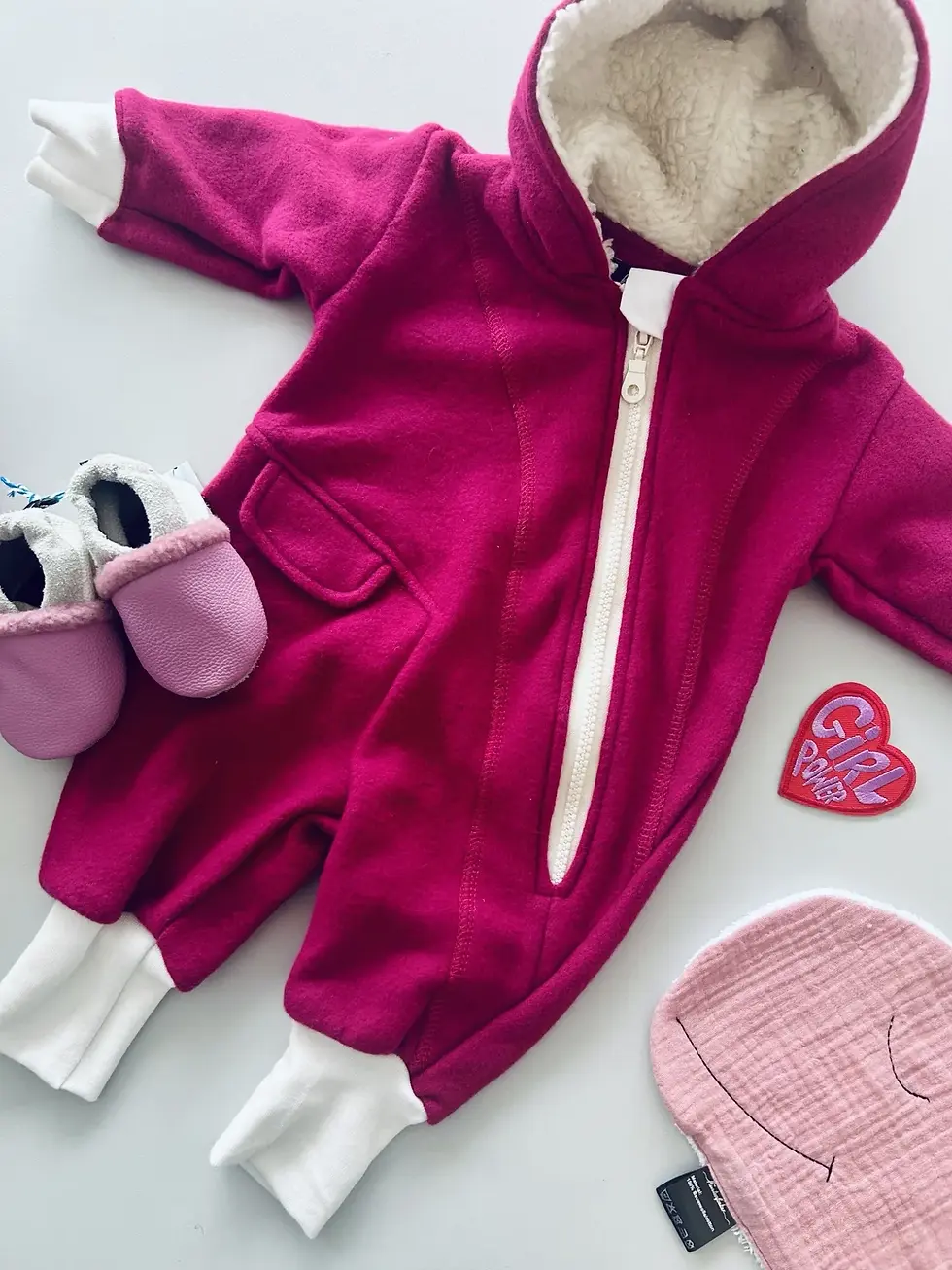 Miniaturbild: Beerenfarbener Bio-Fleeceoverall mit Kapuze, hellem Futter und Reißverschluss liegt neben rosa Babyschuhen und einer bestickt
