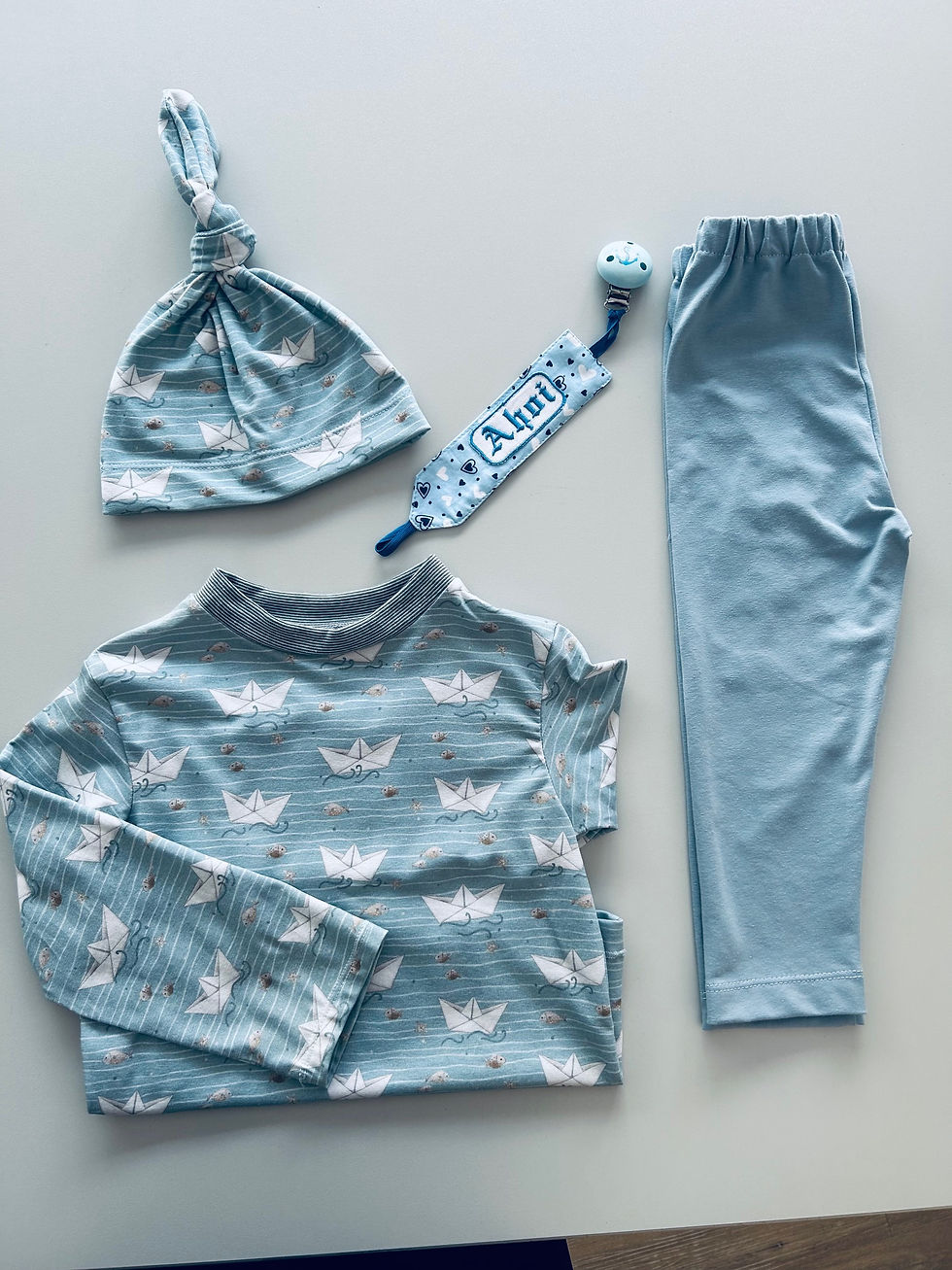 Miniaturbild: Knotenmütze und Langarmshirt in hellblau mit kleinen Papierbooten und Fischen, Schnullerband in hellblau und Kinder Leggings