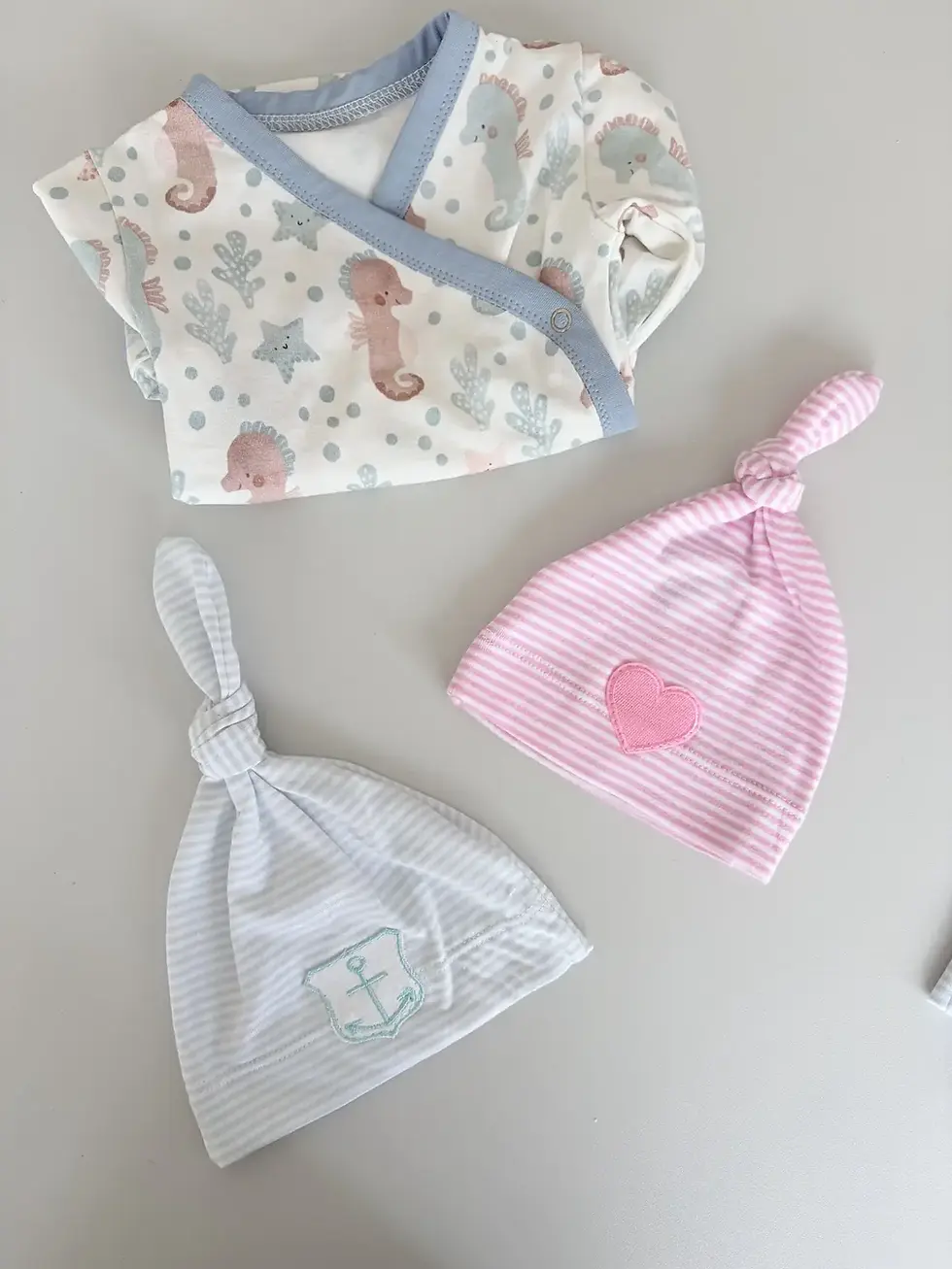 Miniaturbild: Hellblau gestreifte Anker-Knotenmütze und rosa Herz-Knotenmütze neben einem maritimen Baby-Wickelbody.