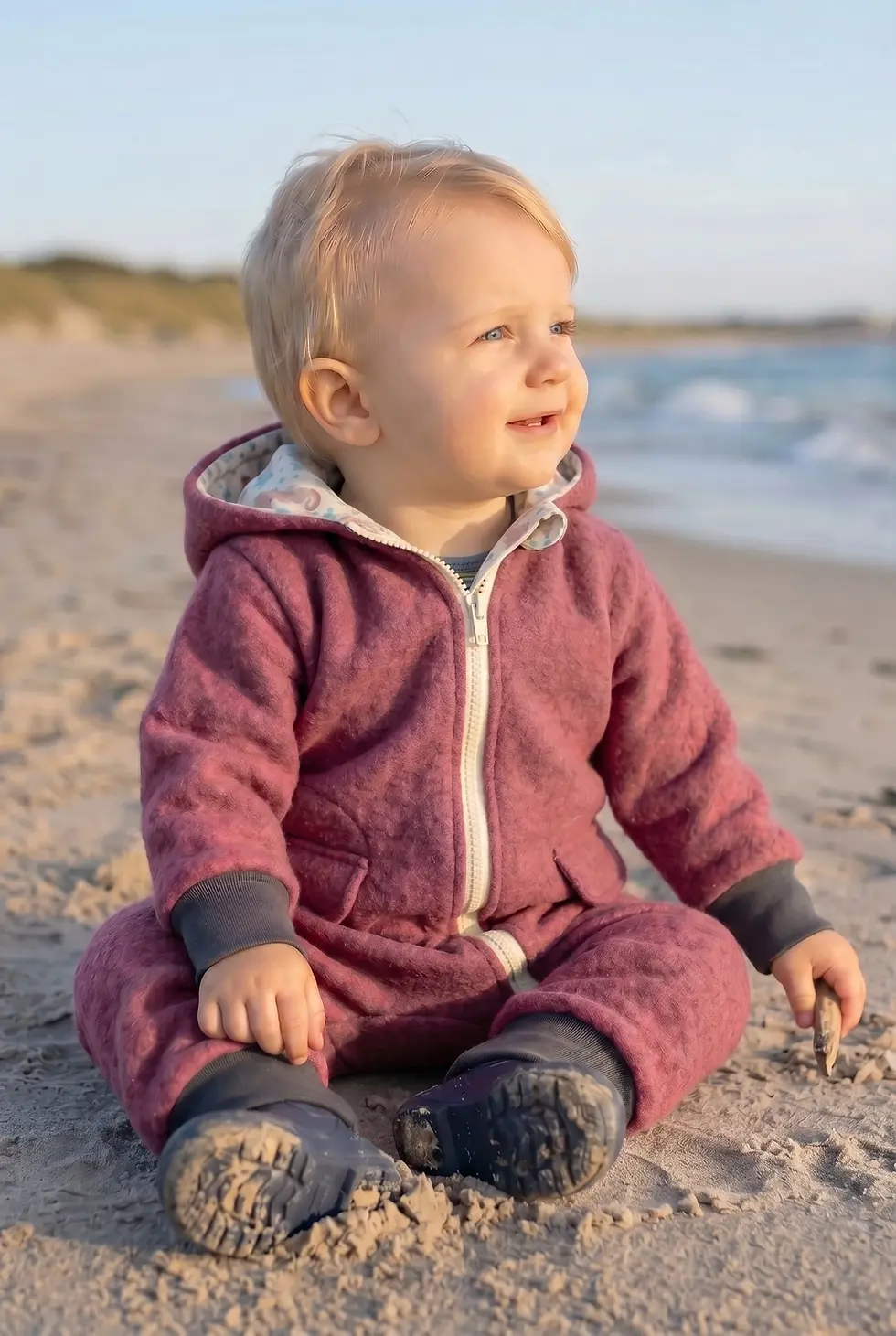 Miniaturbild: Baby trägt den Walkoverall „Zarte Seepferdchen“ aus Wollwalk in Beerenrosa am Strand bei kühlem Wetter.