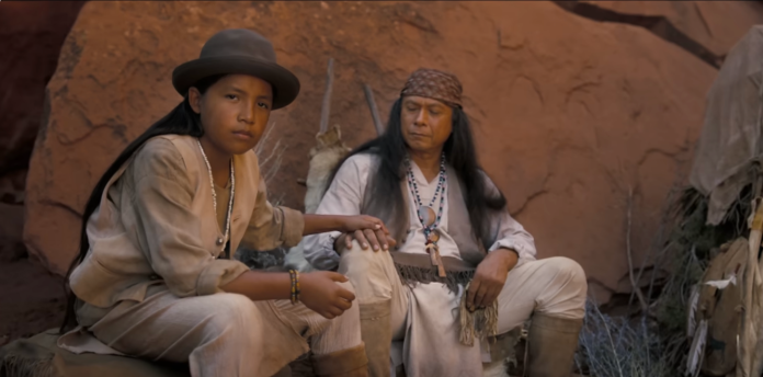 two apaches sit on a rock against a rock formation behind them. the boy on the left wears a bowler hat, loose white shirt and trousers. he has his hand on the older man's hand. the old man is the elder of the tribe and wears a headband, loose white shirt, vest, a neckpiece with beads around his neck and moccasin boots while looking at the young man
