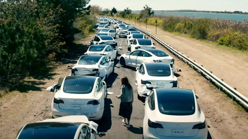 A woman looks at a road with two lanes of Tesla white vehicles that have crashed into the rear ends of all the other cars.  There are dozens of crashed and damaged cars.