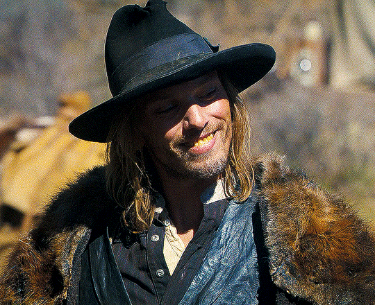 head and shoulders shot of a young man in a wide brimmed hat who us smiling and wearing a fur coat and vest. his hair is long and straggly.