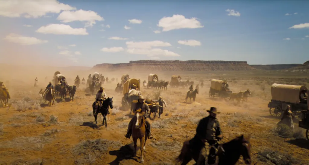 a wide shot of cowboys and settlers on horses and in wagons crossing a wide prairie and arising a lot of dry dust with mountains in the distance