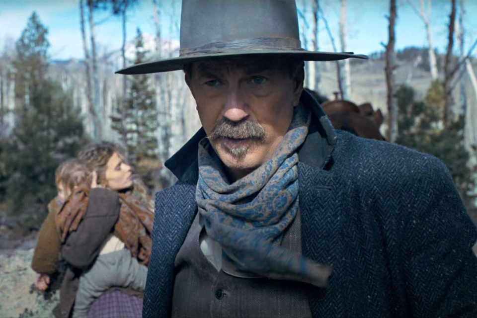 head and shoulder shot of a cowboy in a wide brimmed hat, large moustache, paisley kerchief around his neck and a heavy coat with a young woman holding a baby in the background with trees in the background
