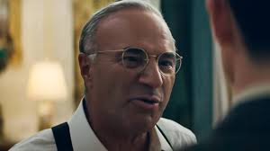 An older man with spectacles and grey hair is speaking sternly to someone.