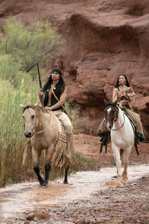 a young Native American apache couple on horseback on white and tan horses in traditional apache clothing ride through a creek in a canyon with the walls of the canyon behind them
