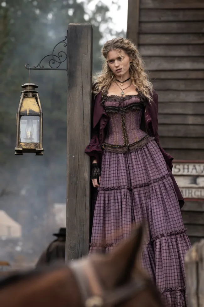 a young woman wearing a purple corset long dress leans against a post in an old west town.  she is a prostitute looking to attract attention of cowboys who have entered the town