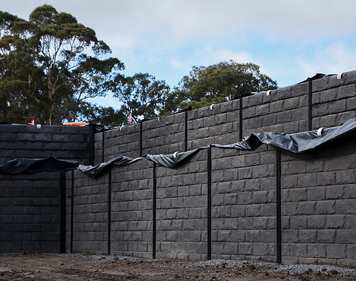 Charcoal concrete sleeper retaining wall with a block-style pattern, supported by black steel posts. The wall is partially covered with black plastic sheeting along the top edge, indicating ongoing installation. Bare soil and gravel are in the foreground, with tall trees in the background under a cloudy sky. Ideal example of a durable retaining wall system using charcoal concrete sleepers and steel post construction.