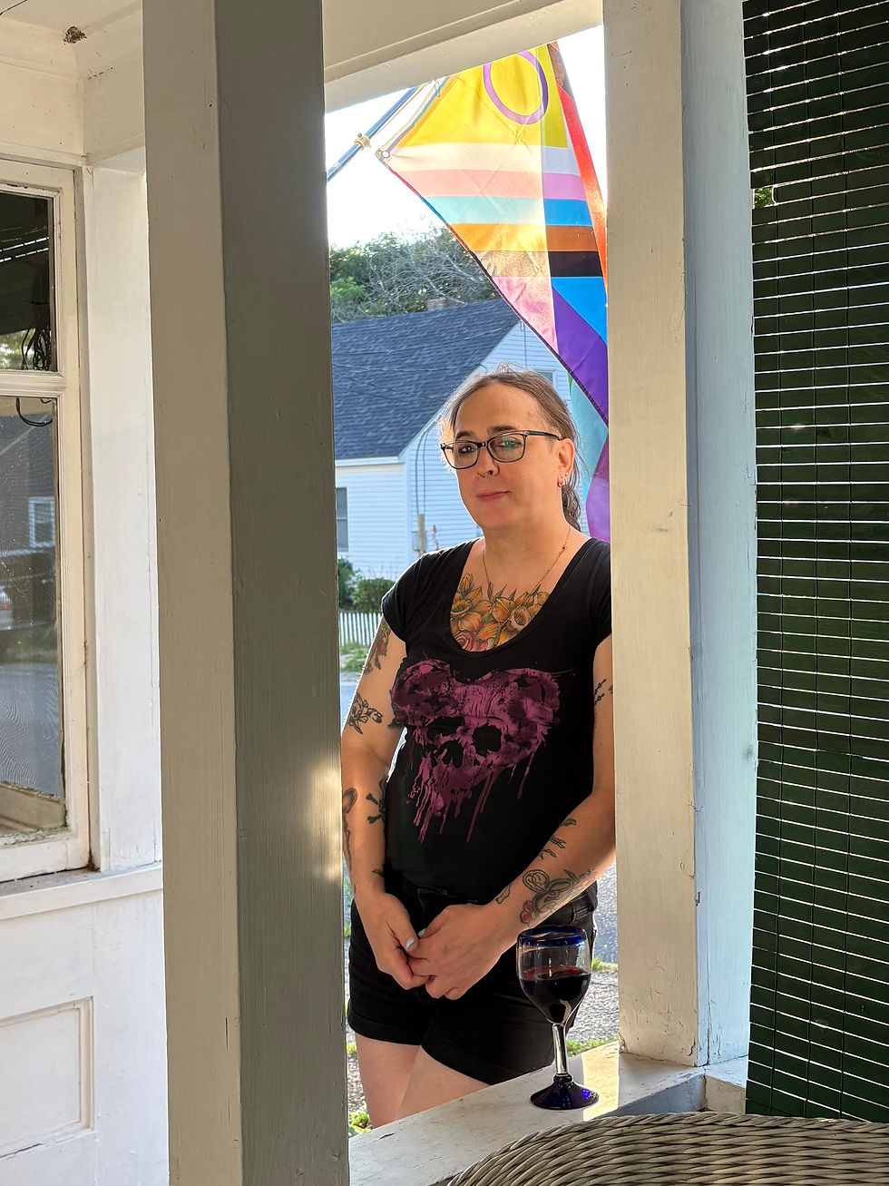 Portrait photo of Billie standing on a porch with a Progress Pride flag in the background, vertical orientation.