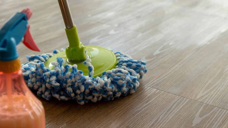 Blue mop and orange spray bottle on wooden floor, cleaning setting. Mop has green handle and blue fibers, conveying tidiness.